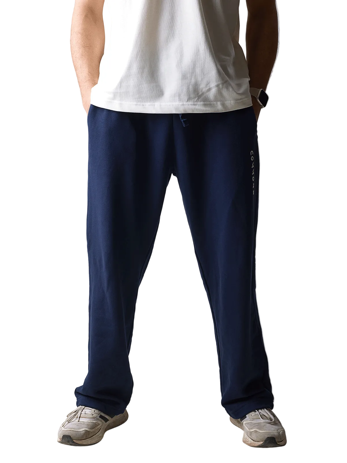 A person wearing navy blue sweatpants with a vertical white logo on the side, paired with a white t-shirt and beige sneakers, standing against a light gray studio background.