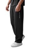A person wearing black wide-leg sweatpants with vertical white 'CANYONS' branding on the left leg, paired with white sneakers, photographed against a light gray studio background.
