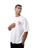 A man wearing a white oversized t-shirt with a colorful flower graphic and the word 'CAMPING' printed on the chest, posed against a solid gray studio background.
