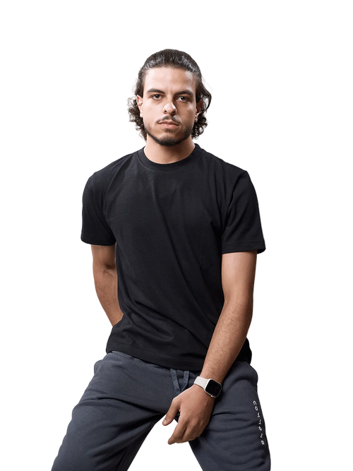 A man wearing a black short-sleeve crewneck t-shirt and dark gray sweatpants, sitting against a light gray studio background.