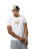 A man wearing a white t-shirt with yellow-green trim and a 'NERD' graphic on the chest, paired with a light beige cap and light blue jeans, standing against a solid gray background.