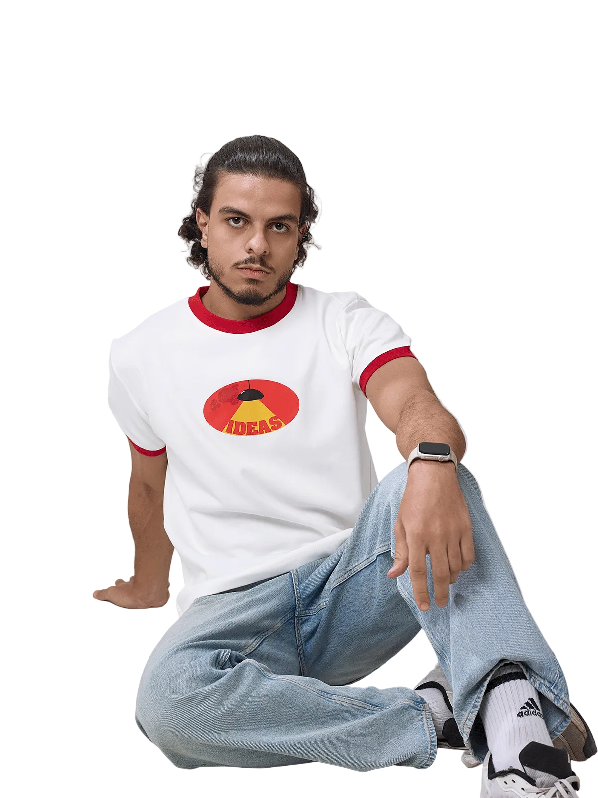 A man sitting on a light gray floor wearing a white ringer t-shirt with red trim and a circular graphic on the chest, paired with light blue jeans, white socks, and black sneakers.