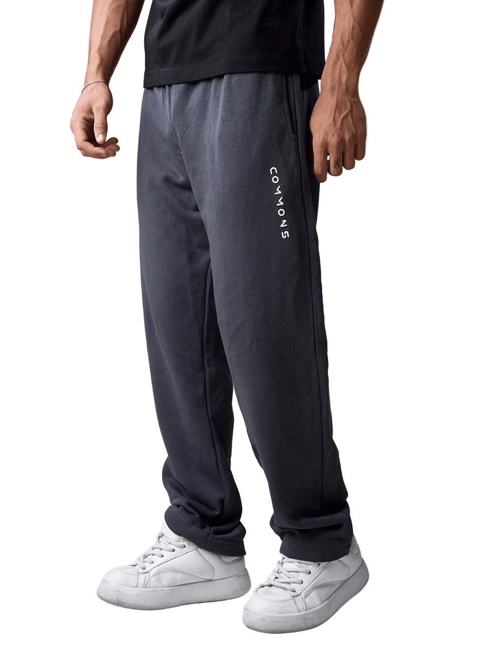 A person wearing dark charcoal gray sweatpants with vertical white 'COMMON' text on the left leg, paired with a black t-shirt and white sneakers, shown from the waist down against a light gray studio background.