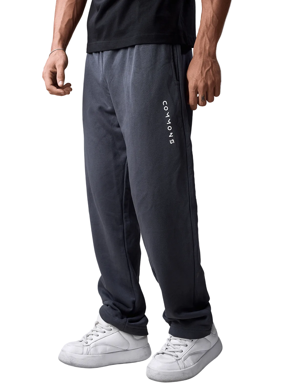 A person wearing dark charcoal gray sweatpants with vertical white 'COMMON' text on the left leg, paired with a black t-shirt and white sneakers, shown from the waist down against a light gray studio background.