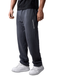 A person wearing dark charcoal gray sweatpants with vertical white 'COMMON' text on the left leg, paired with a black t-shirt and white sneakers, shown from the waist down against a light gray studio background.