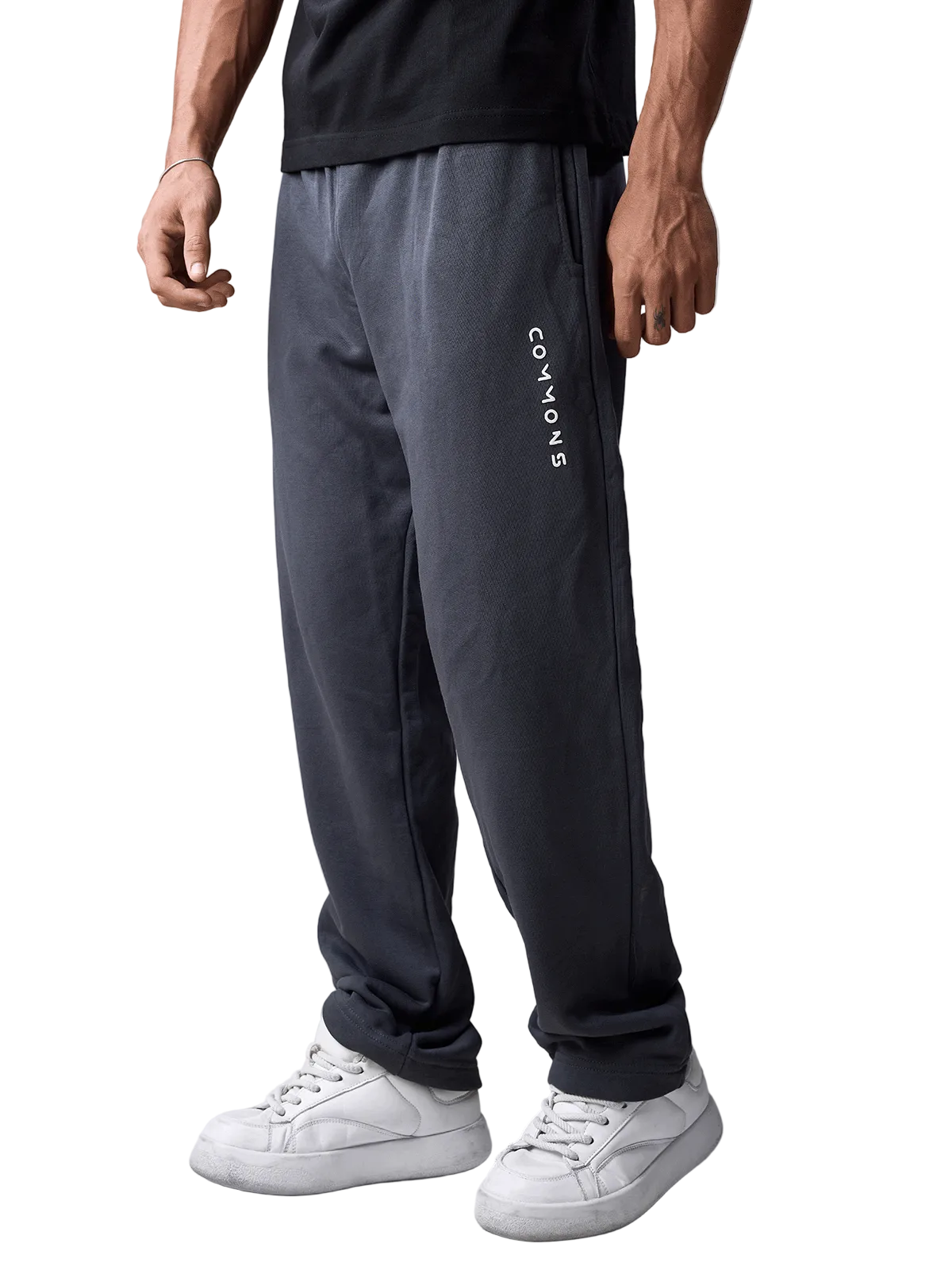 A person wearing dark charcoal gray sweatpants with vertical white 'COMMON' text on the left leg, paired with a black t-shirt and white sneakers, shown from the waist down against a light gray studio background.