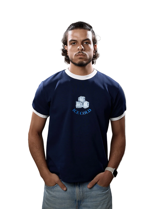 A man wearing a navy blue ringer t-shirt with white trim and a graphic of ice cubes and the text 'ICE COLD' on the chest, paired with light blue jeans, standing against a solid gray background.