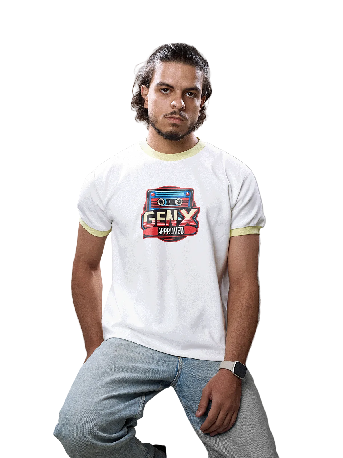A man wearing a white ringer t-shirt with light yellow trim and a 'GENX APPROVED' cassette graphic on the chest, paired with light blue jeans, sitting on a wooden stool against a gray studio background.