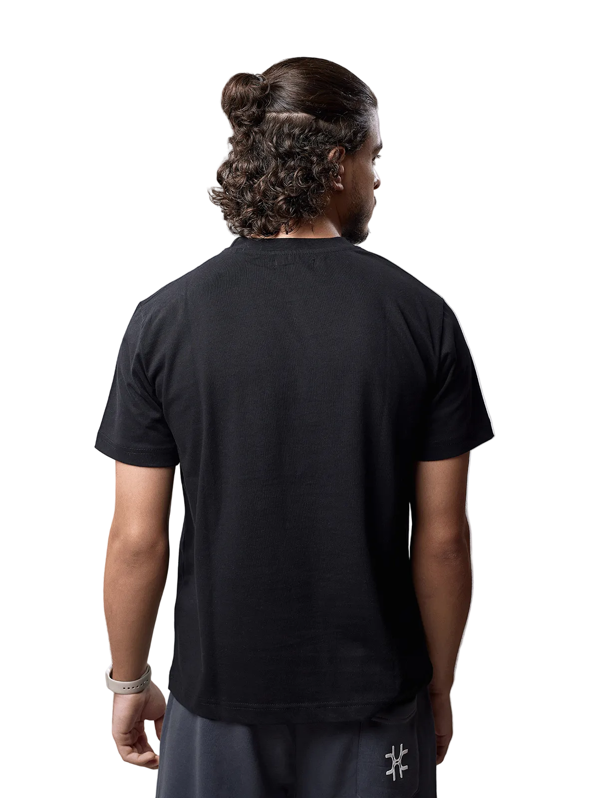 A man viewed from behind wearing a black short-sleeve crewneck t-shirt and dark gray pants, standing against a light gray studio background.