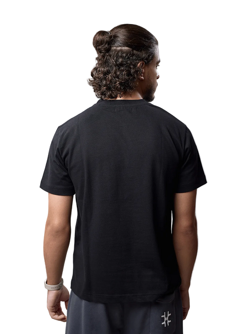 A man viewed from behind wearing a black short-sleeve crewneck t-shirt and dark gray pants, standing against a light gray studio background.