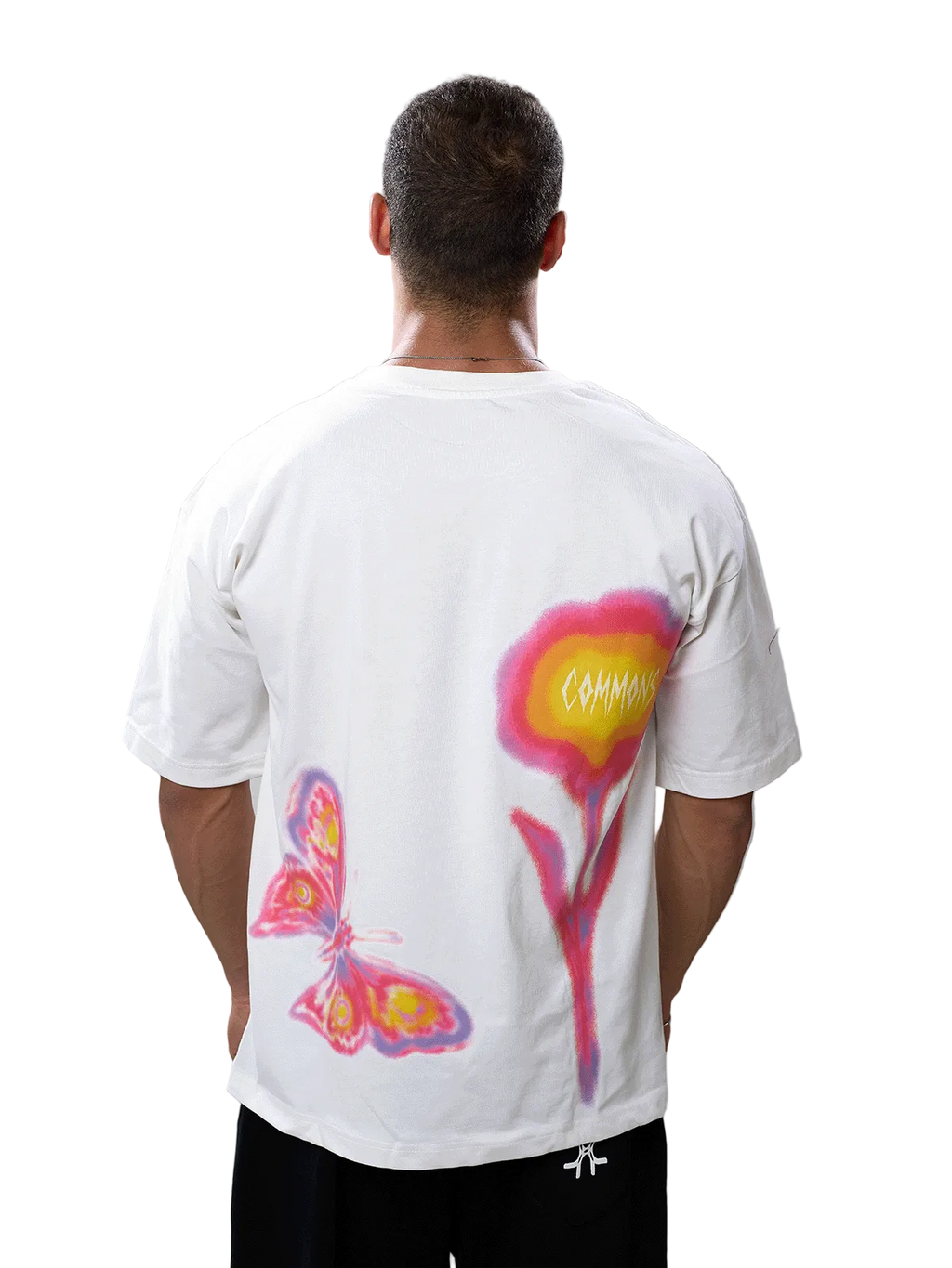 A man viewed from behind wearing a white t-shirt with a large graphic print on the back featuring a pink and yellow flower and a butterfly, paired with black pants against a gray studio background.