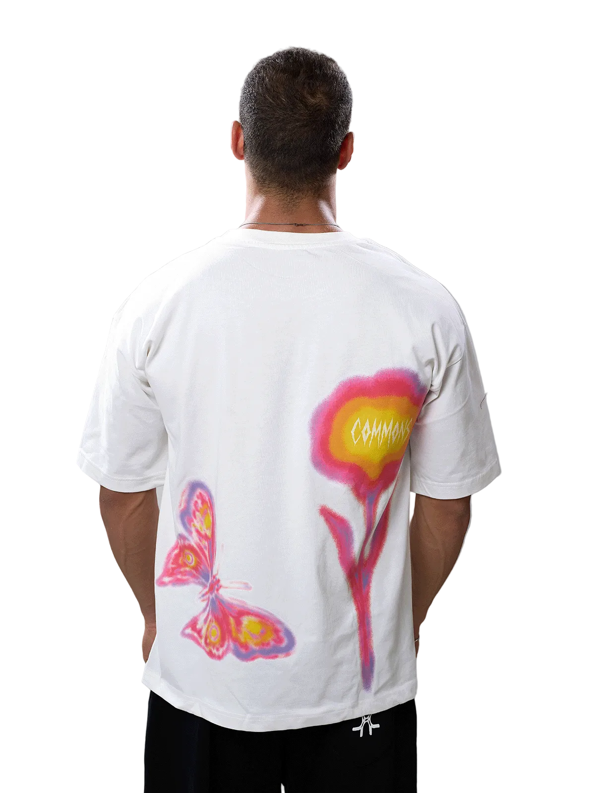 A man viewed from behind wearing a white t-shirt with a large graphic print on the back featuring a pink and yellow flower and a butterfly, paired with black pants against a gray studio background.