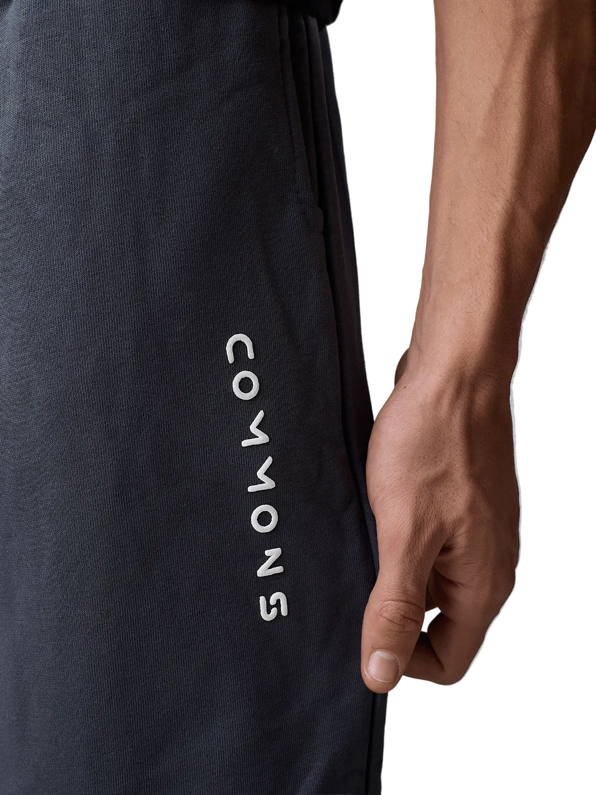Close-up of a person wearing dark charcoal gray athletic sweatpants with the brand name 'COMMONS' printed vertically in white on the side, with the model's hand resting on the thigh.