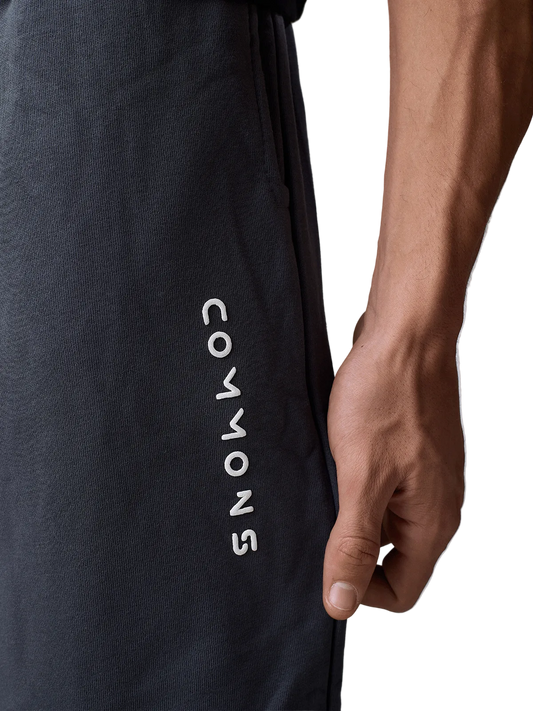 Close-up of a person wearing dark charcoal gray athletic sweatpants with the brand name 'COMMONS' printed vertically in white on the side, with the model's hand resting on the thigh.