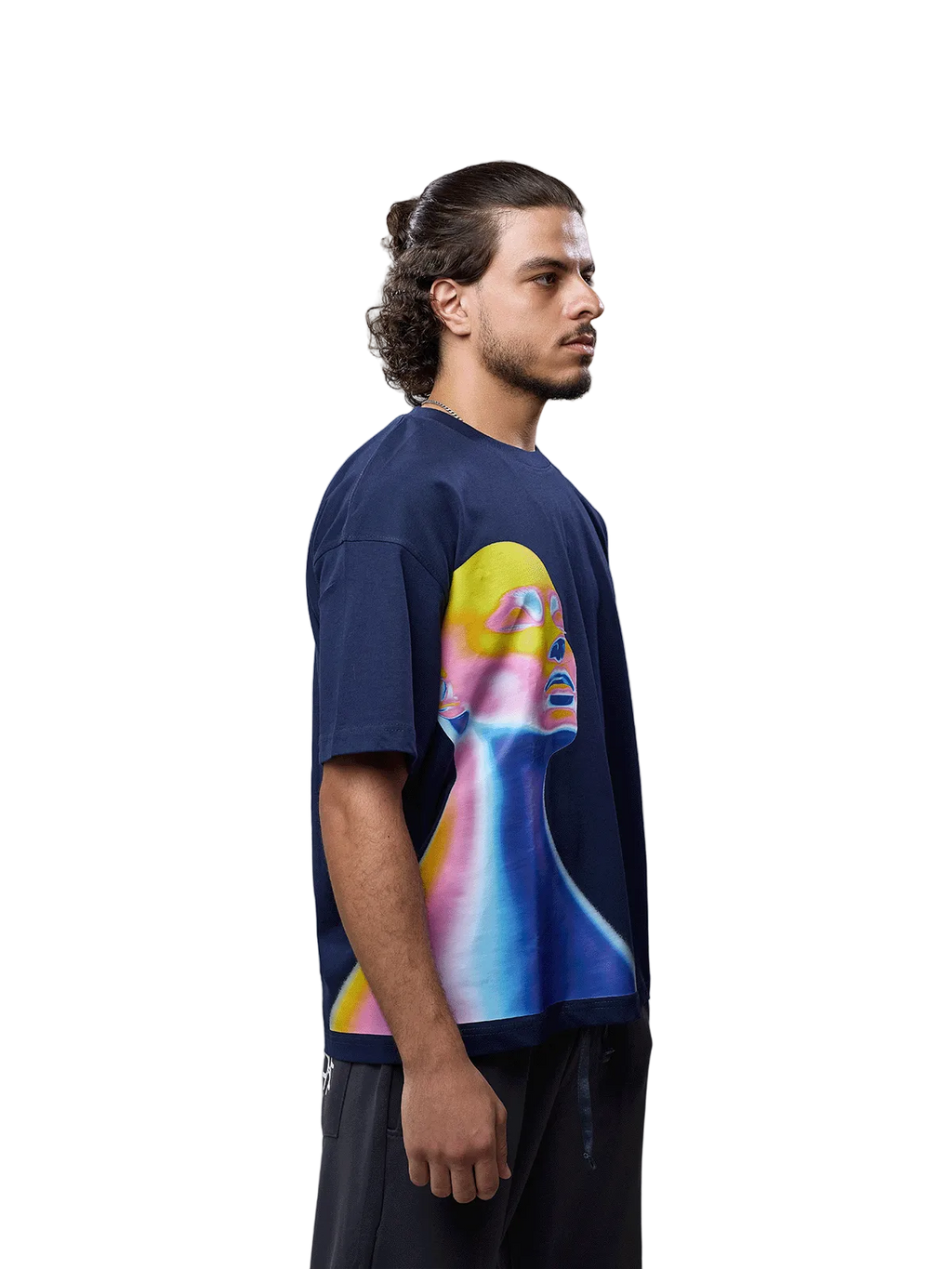 A man wearing a navy blue oversized t-shirt with a large, colorful graphic print of a stylized face on the front, paired with dark pants, photographed against a neutral gray studio background.