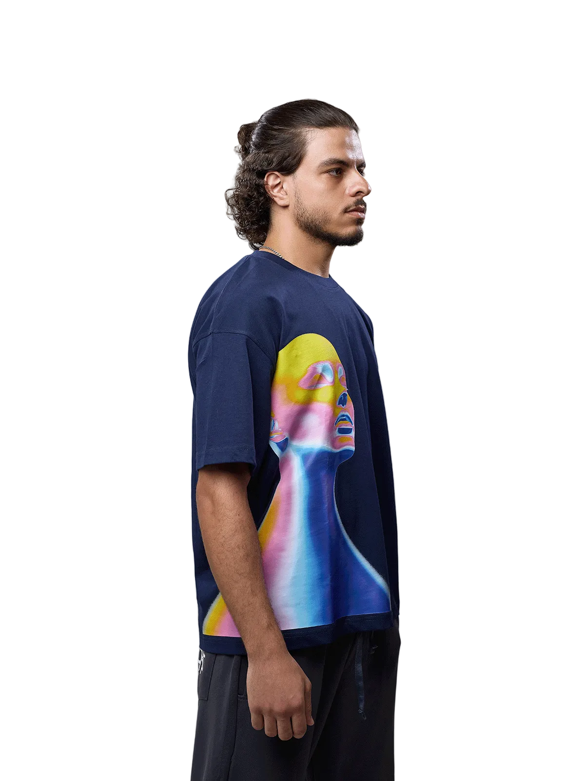 A man wearing a navy blue oversized t-shirt with a large, colorful graphic print of a stylized face on the front, paired with dark pants, photographed against a neutral gray studio background.