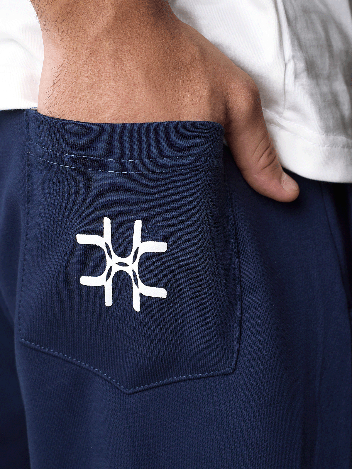 Close-up of a person's hand in a white shirt pocketing navy blue sweatpants with a white embroidered geometric logo on the back pocket.