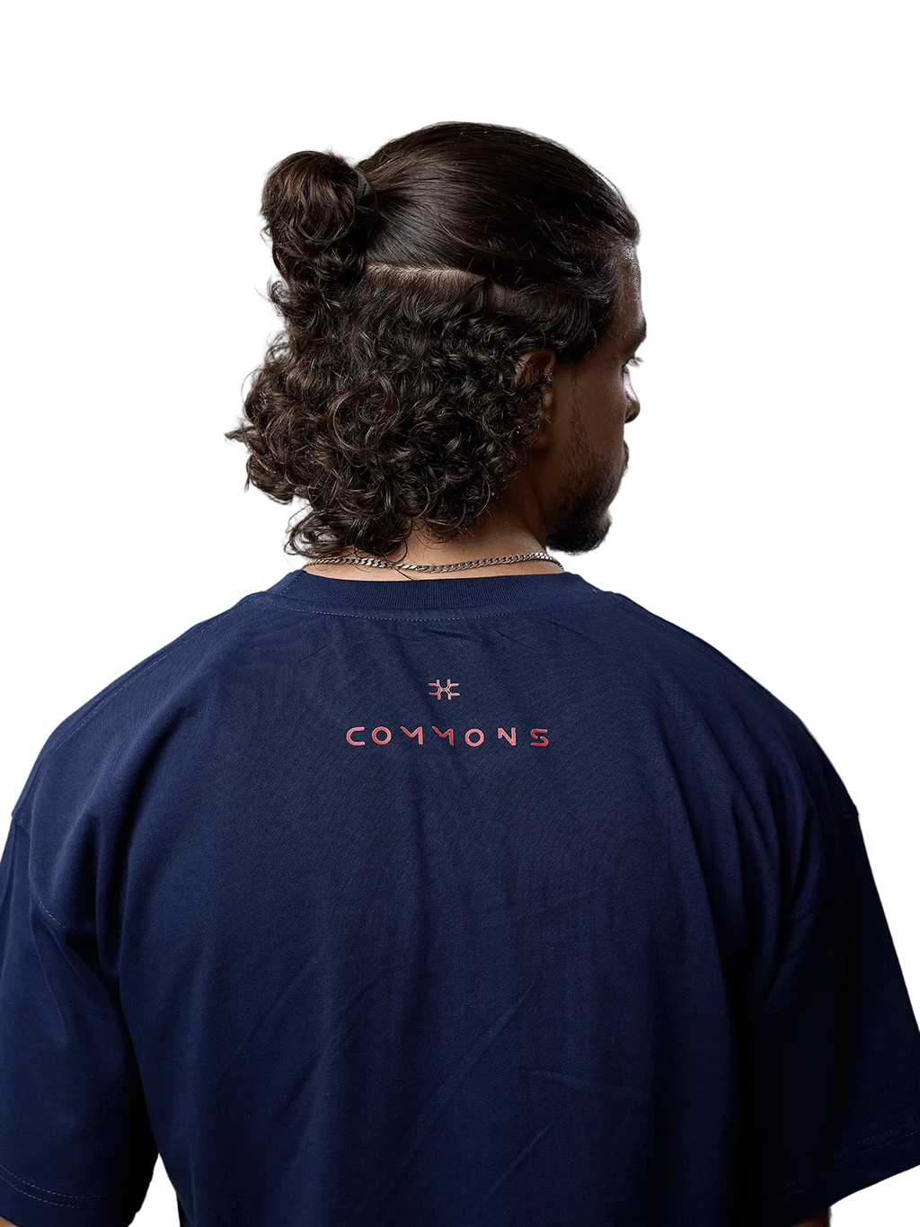 A man viewed from behind wearing a navy blue t-shirt with 'COMMONS' and a star logo printed in orange on the back, set against a plain light gray background.