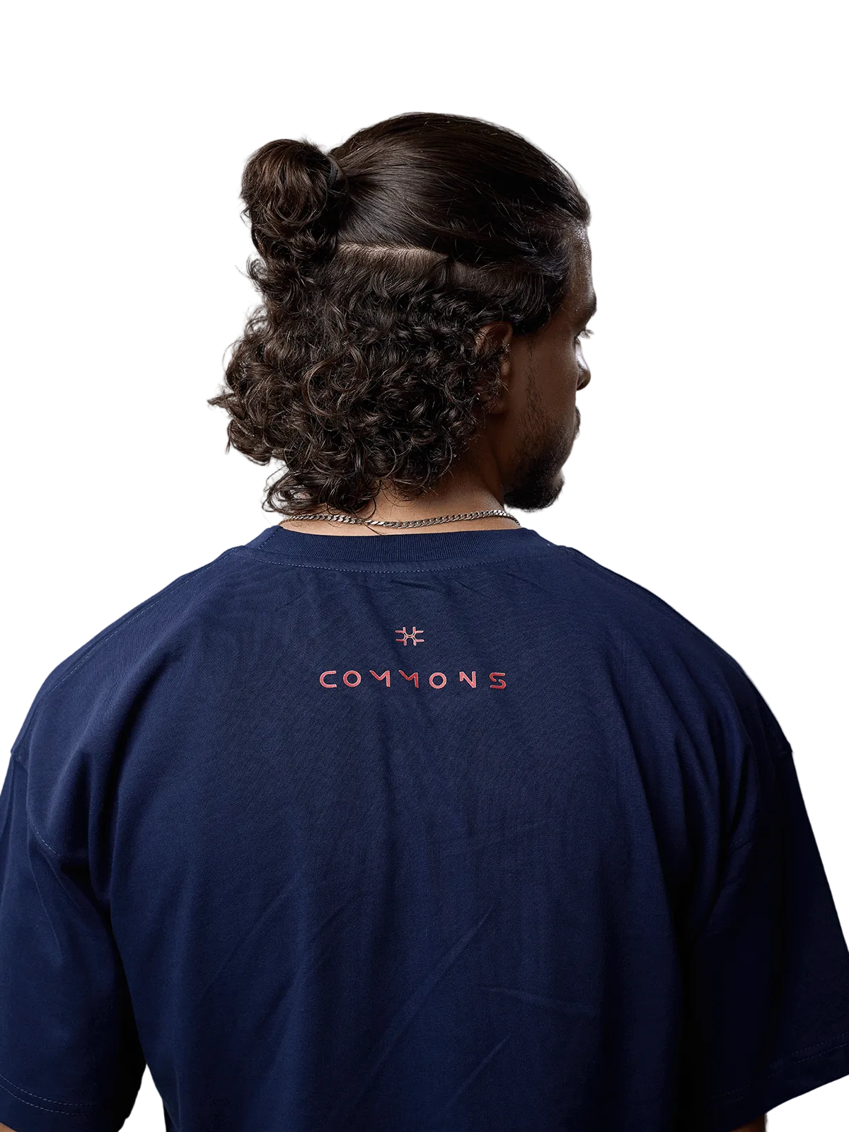 A man viewed from behind wearing a navy blue t-shirt with 'COMMONS' and a star logo printed in orange on the back, set against a plain light gray background.
